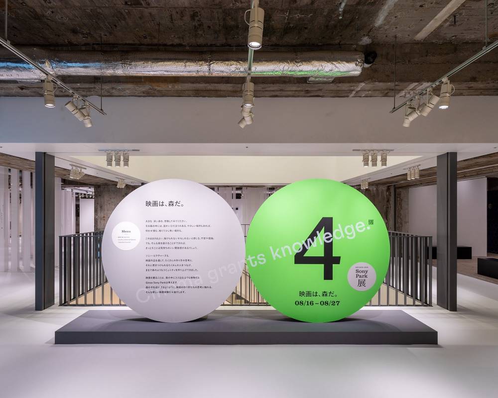 Exhibit. A round gray art object with the exhibition statement, and a green version of the same object labeled “4,” are displayed side by side at the entrance to the venue on the second basement floor.