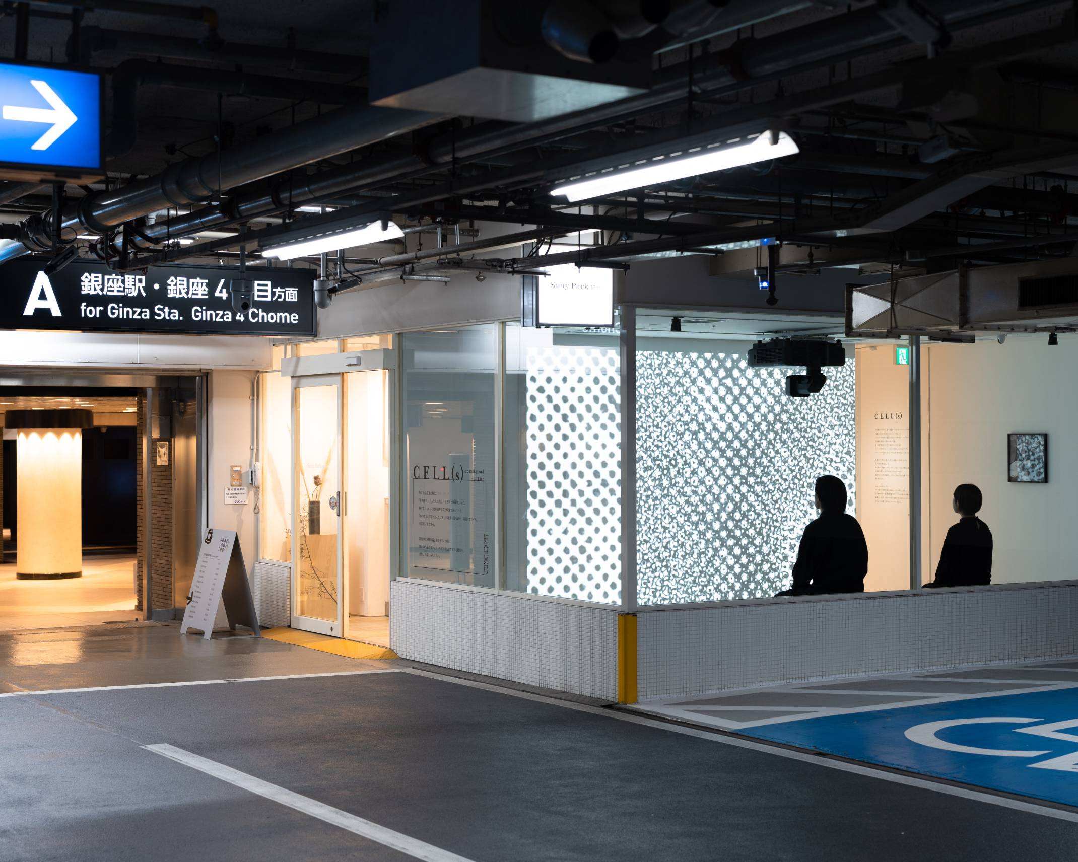 Exterior photo. Two visitors sit and gaze at the art piece projected onto the wall inside of the exhibition space.
