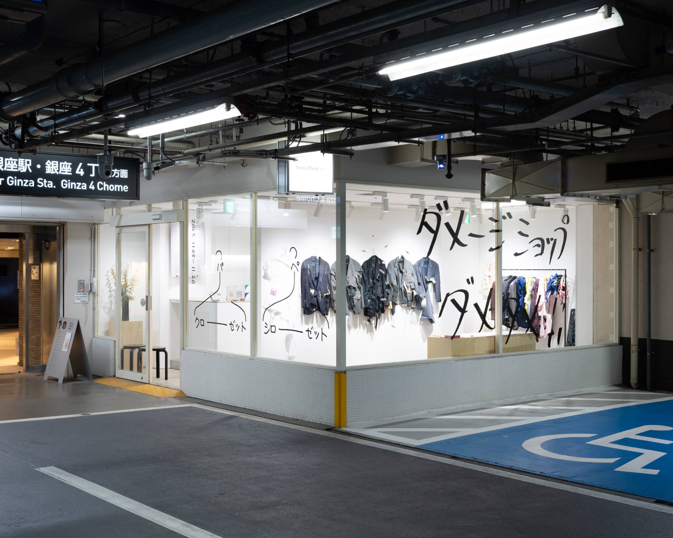 Exterior. The glass window of the exhibition space, which features rows of clothing items cut in a variety of ways, has the words “Damage Shop No No!!” written in large letters in Japanese.