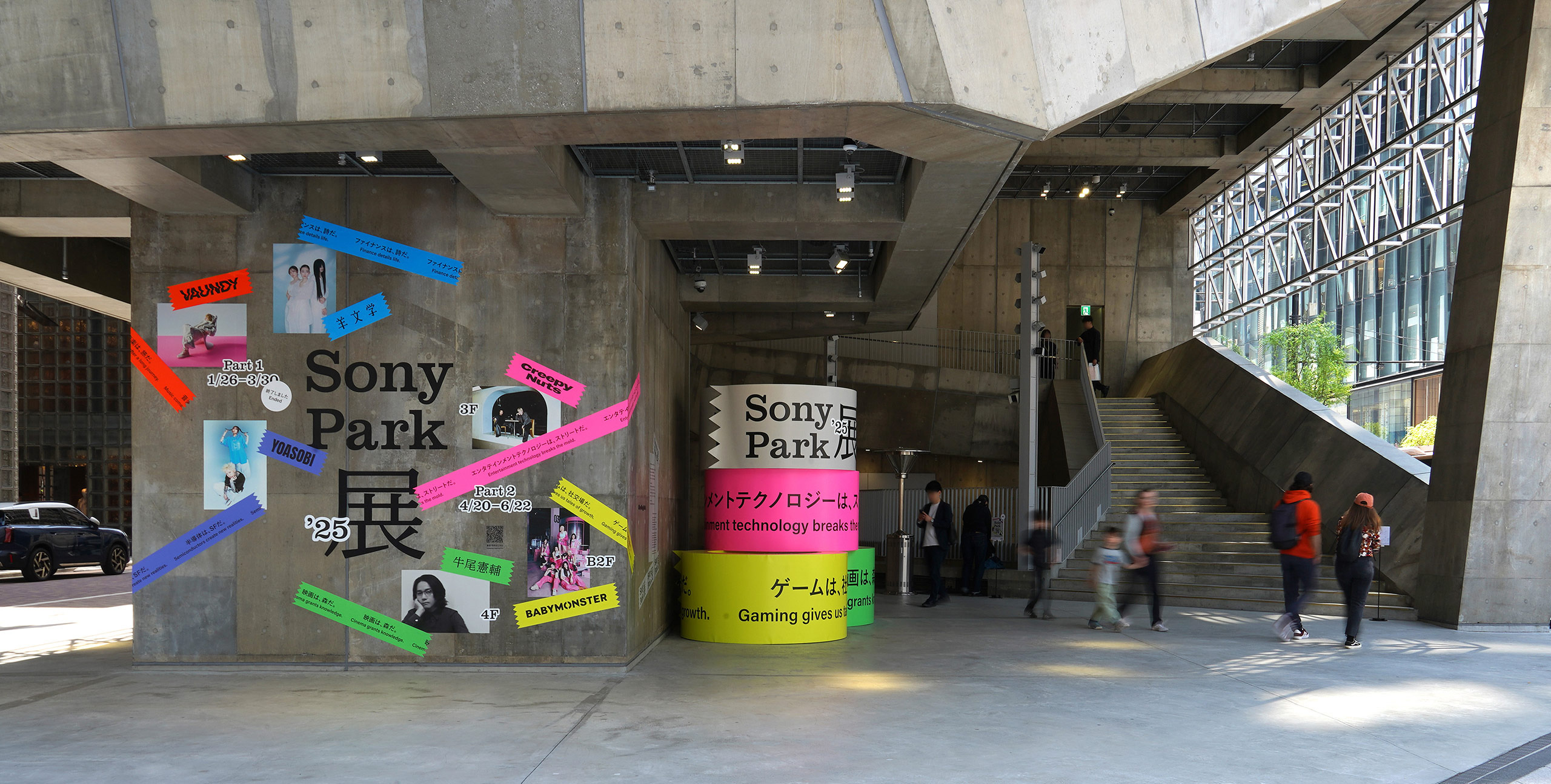 Entrance of Ginza Sony Park. A concrete wall is decorated with colorful banners and posters featuring artist names for the “Sony Park Exhibition.” On the right, people are seen walking up the stairs.