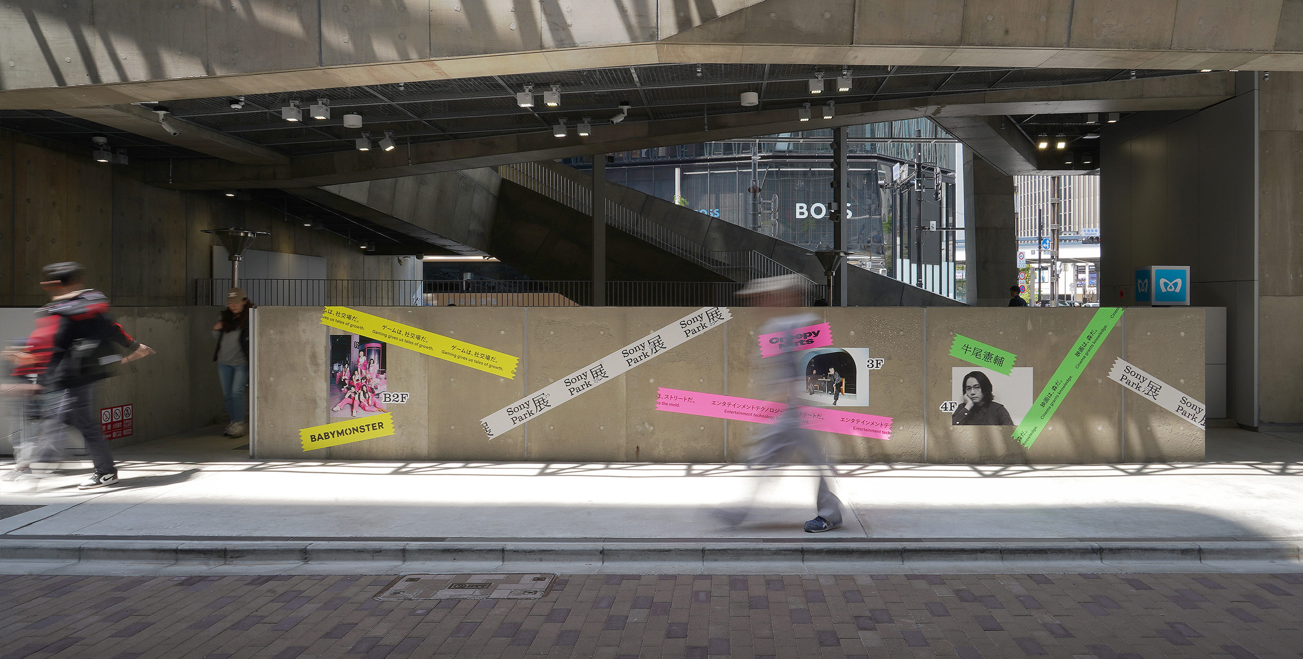 Street-level view of Ginza Sony Park. A concrete wall displays the “Sony Park Exhibition” logo and visuals of participating artists, with pedestrians walking along the sidewalk.